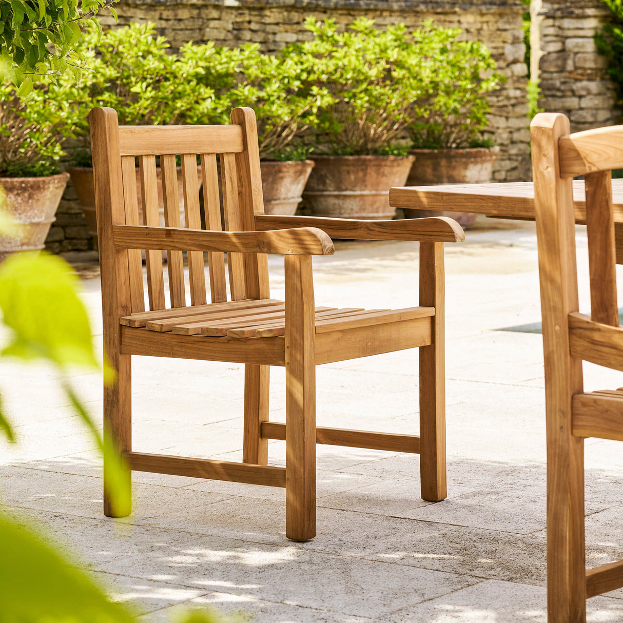 Wooden outdoor chairs on a stone patio with greenery in the background